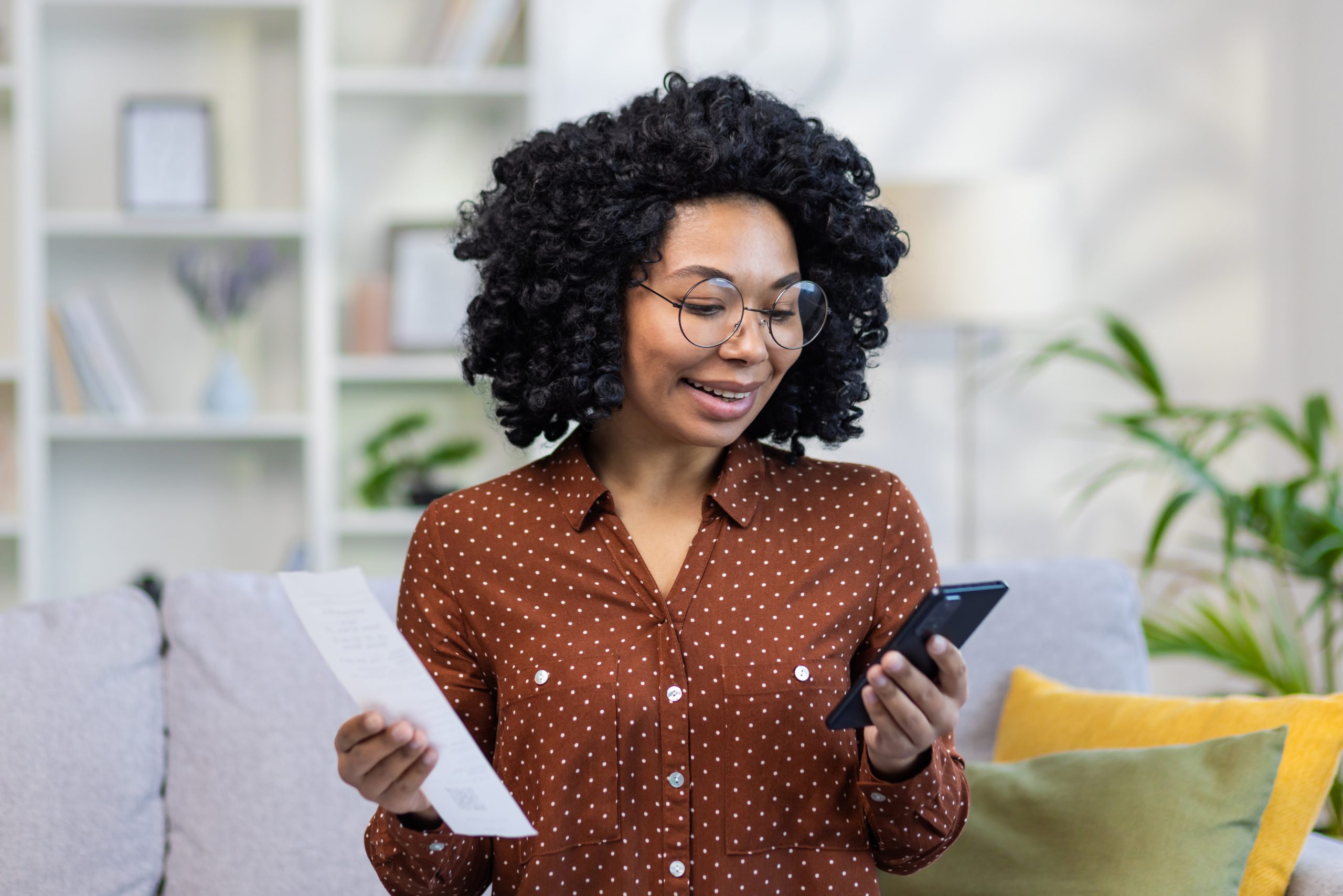 Closeup photo of a smiling young African-American woman in glasses sitting on the sofa at home, holding documents and bills, using a mobile phone